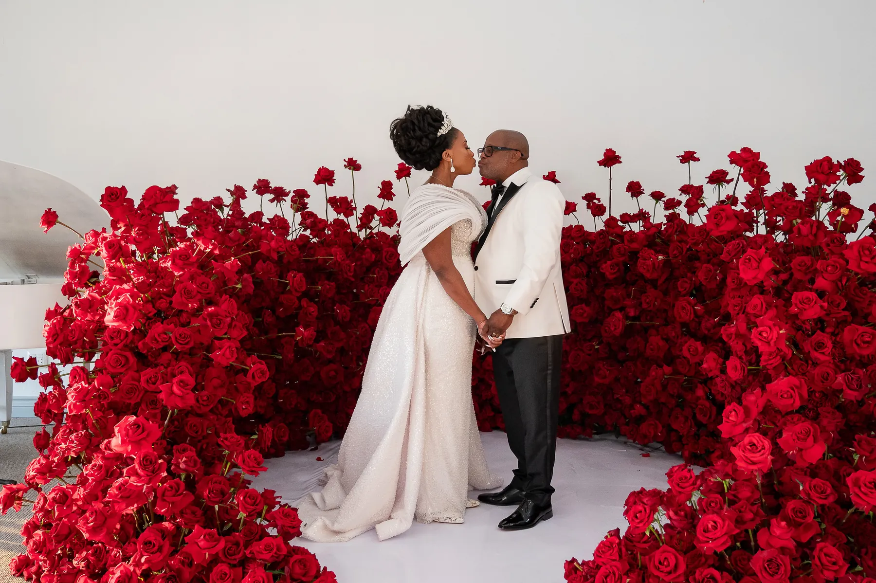Bride and groom kissing, framed by a dense semicircle of red roses against a white backdrop
